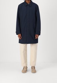 Persoon in een marineblauwe lange jas, beige broek en beige veterschoenen, staande tegen een effen lichte achtergrond.