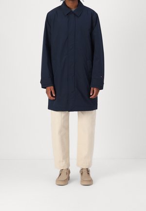 Personne portant un long manteau bleu marine, un pantalon beige et des chaussures en cuir beige, se tenant contre un fond clair.