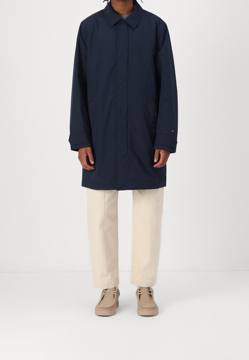 Persoon in een marineblauwe lange jas, beige broek en beige veterschoenen, staande tegen een effen lichte achtergrond.