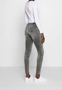 Mujer vistiendo una blusa blanca de manga larga, jeans ajustados gris desgastados y zapatillas blancas, de pie con la espalda ligeramente girada.