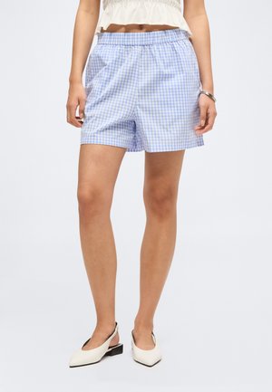 Vrouw die blauw-witte geruite shorts draagt, een wit cropped topje, witte slingback hakken, staat met handen ontspannen langs haar zij.