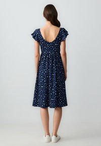 Navyblaues Blumenkleid mit kurzen Rüschensäumen, einem offenen Rücken und einem taillierten Oberteil, das in einen knielangen Rock übergeht. Kombiniert mit weißen Sneakers.