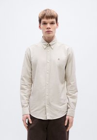 Ανοιχτό μπεζ πουκάμισο από βαμβάκι με κουμπιά, που διαθέτει μυτερό γιακά, μακριά μανίκια και διακριτική λεπτομέρεια λογότυπου στο στήθος. Λείο ύφασμα.