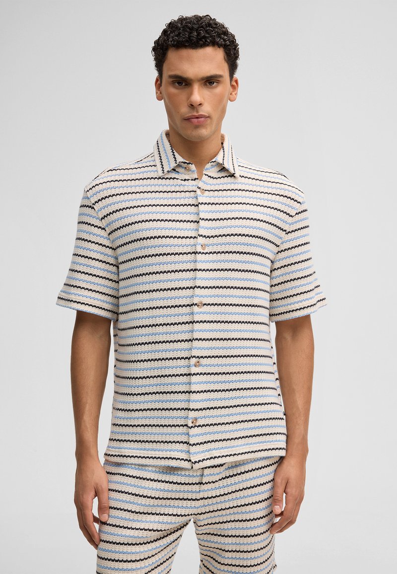 Homme portant une chemise à manches courtes boutonnée et un pantalon assorti avec des rayures ondulées horizontales bleues, noires et beiges sur un fond clair.