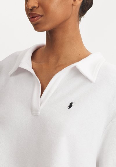 Valkoinen polo-paita, joka on valmistettu pehmeästä kankaasta, jossa on kaulus ja pieni musta logo rinnassa. Lähikuva näyttää kankaan rakenteen ja ompelu yksityiskohdat.
