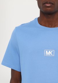 Lyseblå bomulls T-skjorte med rund hals og korte ermer, med et hvitt brodert "MK"-logo innenfor en rektangulær kontur.