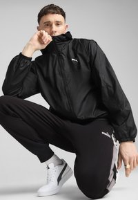 Sort letvægtsjakke med høj krave og elastiske manchetter, parret med sorte joggers med en stribe og hvide sneakers.