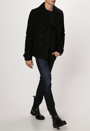 Sort dobbeltradet peacoat med brede revers, båret over en sort skjorte, kombineret med mørke jeans og sorte ankelstøvler med nitter.