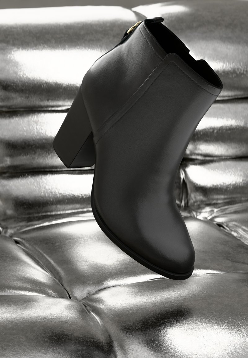 Bottine en cuir noir avec un talon carré, surface lisse, bout légèrement pointu et accent de fermeture éclair dorée. Fond métallique.