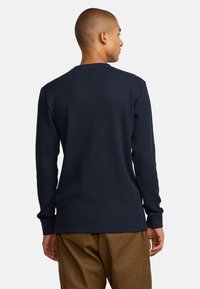Marinefarbener langärmliger, gerippter Thermo-Pullover mit Rundhalsausschnitt und enganliegenden Bündchen, aus strukturiertem Stoff; getragen mit braunen Hosen.