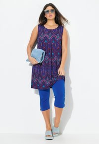 Ärmellose Tunika in Lila mit geometrischem Muster, kombiniert mit blauen Capri-Leggings und hellblauen Rutsch-Sandalen, während sie einen Denim-Artikel hält.