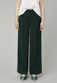Pantalons larges, de couleur vert foncé, en tissu lisse, avec des plis sur le devant et une taille cintrée avec des poches latérales. Associés à des baskets décontractées.