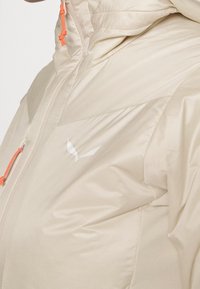 Beige eristetty takki, jossa on sileä pinta, varustettu vetoketjulla ja oransseilla naruilla. Valkoinen logo rinnassa, korkea kaulussuunnittelu.