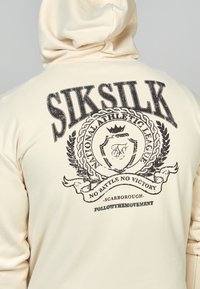 Persona vistiendo una sudadera color crema con el texto negro "SIKSILK" y un emblema atlético que presenta una corona y unaurel en la parte posterior.