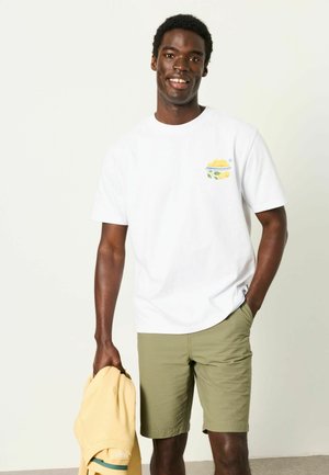 Junger Mann lächelt, trägt ein weißes T-Shirt mit Obstschalen-Design, olivgrüne Shorts, hält in einer Hand eine gelbe Jacke, steht vor einfachem Hintergrund.