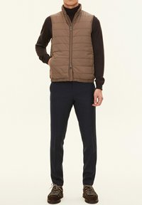 Braune gesteppte Weste mit hohem Kragen und Reißverschluss, getragen über einem dunklen Rollkragenpullover. Kombiniert mit marineblauen, schmal zulaufenden Hosen und braunen Schnürschuhen.