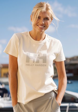 Mujer sonriente con cabello rubio que lleva una camiseta ligera de Helly Hansen y pantalones grises, de pie al aire libre cerca del agua y botes bajo un cielo despejado.