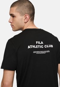 Mustan lyhythihaisen t-paidan takaosassa on valkoinen "FILA ATHLETIC CLUB" -grafiikka, ja se on valmistettu pehmeästä ja sileästä kankaasta.