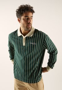 Groene polo met lange mouwen, voorzien van crèmekleurige strepen, een crèmekleurige kraag en een klein geborduurd logo op de borst. Structuurstof, comfortabele pasvorm.