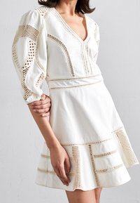Robe blanche avec des manches bouffantes, présentant des accents en crochet, un décolleté en V profond et une jupe à volants avec des découpes horizontales.