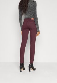 Jeans skinny burdeos con una textura suave, cinturilla alta y bolsillos traseros. Llevados con una camiseta ajustada de manga larga en gris oscuro y tacones altos negros.