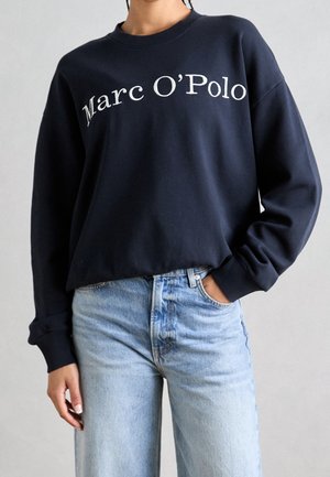 Persoon die een marineblauwe trui met lange mouwen met de tekst "Marc O’Polo" draagt en lichtblauwe hoge taille spijkerbroek, met één hand in de zak.