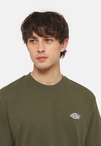 Olivgrön crewneck t-shirt i bomull, med en vit broderad logotyp på vänster bröst och normal passform. Slät textur.