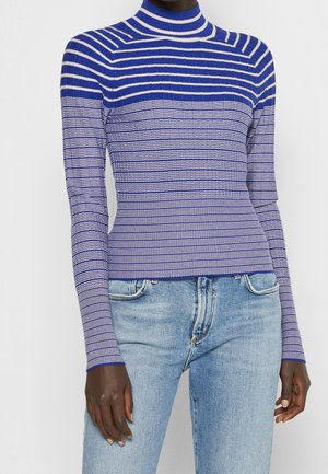 Femme portant un pull à col roulé côtelé bleu et blanc à rayures et un jean en denim bleu clair, debout devant un fond uni.