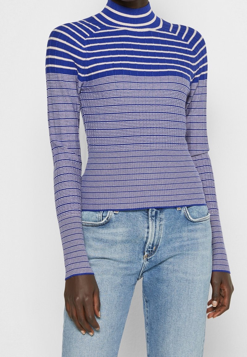 Femme portant un pull à col roulé côtelé bleu et blanc à rayures et un jean en denim bleu clair, debout devant un fond uni.