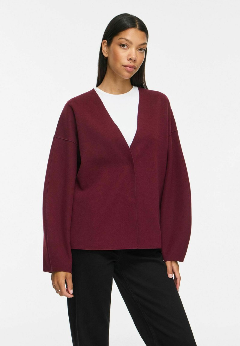 Γυναίκα που φοράει μια άνετη μπορντό ζακέτα πάνω από λευκό τοπ και μαύρο παντελόνι, στέκεται με ουδέτερο βλέμμα μπροστά σε απλό λευκό φόντο.