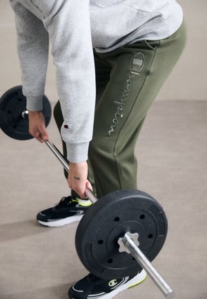 Szara bluza i oliwkowe zielone spodnie dresowe z logo "Champion" wyszywanym. Czarne buty z neonowymi akcentami. Sztanga do podnoszenia ciężarów trzymana obiema rękami.