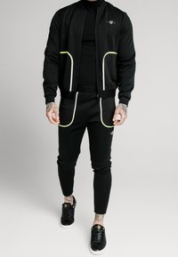 Chándal atlético negro con acentos amarillos reflectantes, que incluye una chaqueta bomber y pantalones ajustados. Completo con zapatillas negras.