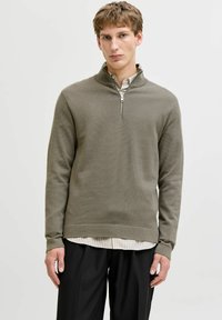 Pull vert olive à col zippé en tissu en maille texturée, porté sur une chemise blanche rayée, associé à un pantalon noir.