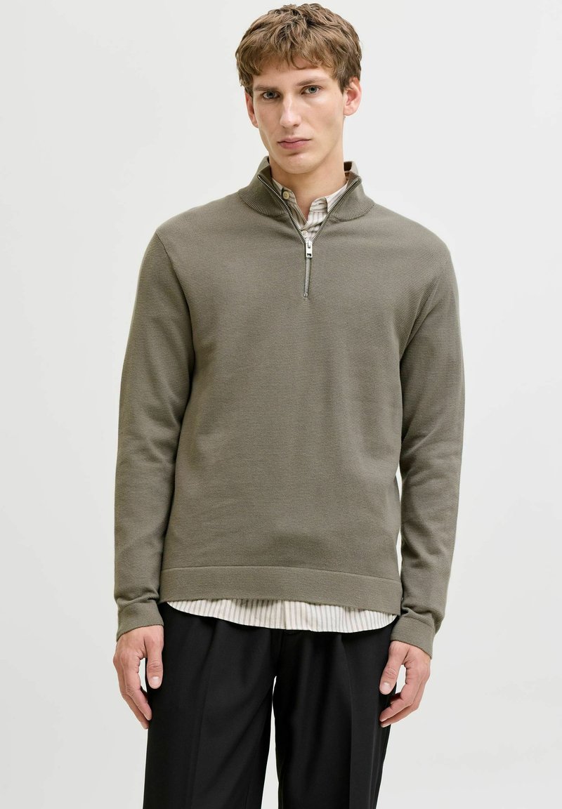 Pull vert olive à col zippé en tissu en maille texturée, porté sur une chemise blanche rayée, associé à un pantalon noir.