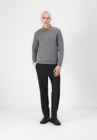 Grauer Pullover mit braunem Kragen und einem 'H'-Logo, kombiniert mit dunklen Hosen und schwarzen Schuhen, vor einem schlichten Hintergrund.