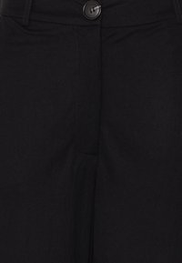 Gros plan sur un pantalon noir avec une taille à boutons et des coutures visibles au niveau de la fermeture éclair.