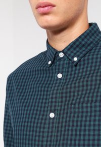 Groen en marineblauw geruit overhemd met knopen en een kraag. Voorzien van witte knopen en een klein zakje aan de linkerkant van de borst.