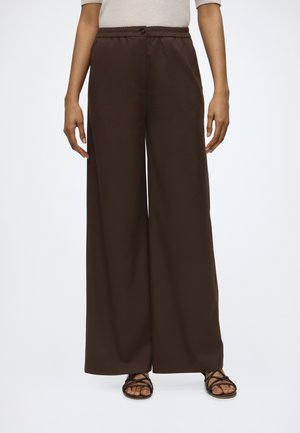 Personne portant un pantalon large marron foncé avec bouton et taille élastique, assorti de sandales noires à brides sur fond blanc.