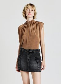 Donna con capelli biondi corti indossa un top marrone senza maniche a collo alto e una mini gonna di jeans nera con cintura e tasche cargo su uno sfondo semplice.