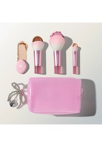 Drie roze make-upborstels met poeder, een roze spons en een roze cosmetische tas met een zilveren hartvormige hanger op een witte ondergrond.