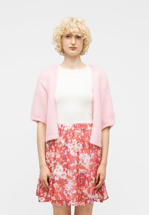 Junge Person mit lockigem blondem Haar, die ein weißes Oberteil, eine rosa kurze Strickjacke und einen roten Blumenrock vor einem schlichten Hintergrund trägt.