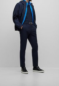 Marinblå vattentät jacka med blå inre foder, tillsammans med en marinblå polo, skräddarsydda marinblå byxor och svarta sneakers med vita sulor.