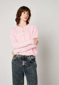 Šviesiai rožinė, pūkuota kardiganas su apvaliu dekolte ir mygtukais priekyje, derinama su tamsiais, nusidėvėjusiais džinsais.