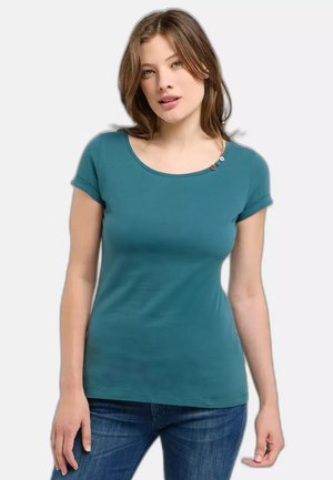 Jeune femme portant un t-shirt ajusté bleu sarcelle à manches courtes avec trois petits boutons près de l'encolure ronde, associé à un jean bleu.