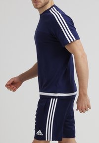 Granatowy komplet sportowy składający się z koszulki z krótkim rękawem oraz spodenek. Koszulka ozdobiona białymi trzema paskami; spodenki mają biały pasek u dołu.