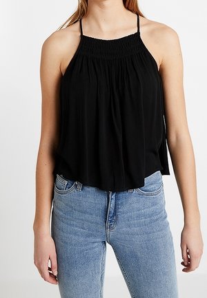 Žena v černém halter topu bez rukávů se shromažďovaným výstřihem a světle modrých džínách s vysokým pasem, stojící před jednobarevným pozadím.