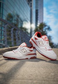 Baskets en cuir blanc avec des accents rouges et une marque. Les caractéristiques comprennent un col rembourré, une semelle texturée et un numéro "550" bien visible sur le côté.