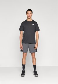 Μαύρο αθλητικό μπλουζάκι με λευκό λογότυπο, συνδυασμένο με γκρι σορτς με πλαϊνές τσέπες. Φορεμένο με μαύρα αθλητικά παπούτσια και κάλτσες.