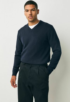 Pulover bleumarin cu decolteu în V, dintr-un material moale, cu mâneci lungi, purtat cu pantaloni negri plisati, model stând în picioare cu o mână în buzunar.