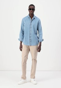 Cămașă din denim albastru deschis cu guler, mâneci lungi, închidere cu nasturi; asortată cu pantaloni bej și adidași albi. Ochelari de soare negri purtați.